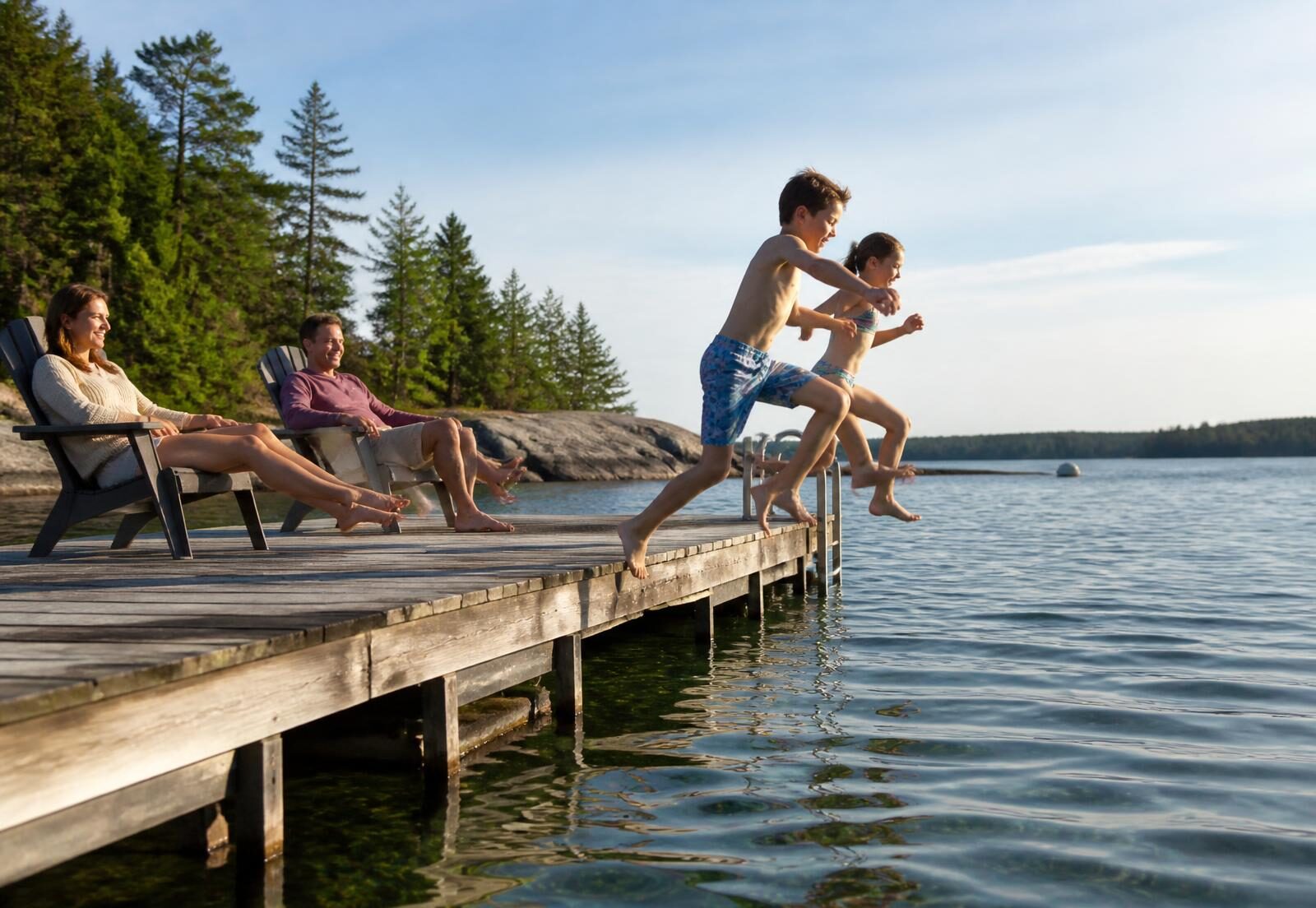 Children jumping off a dock into a lake while parents sit in Adirondack chairs